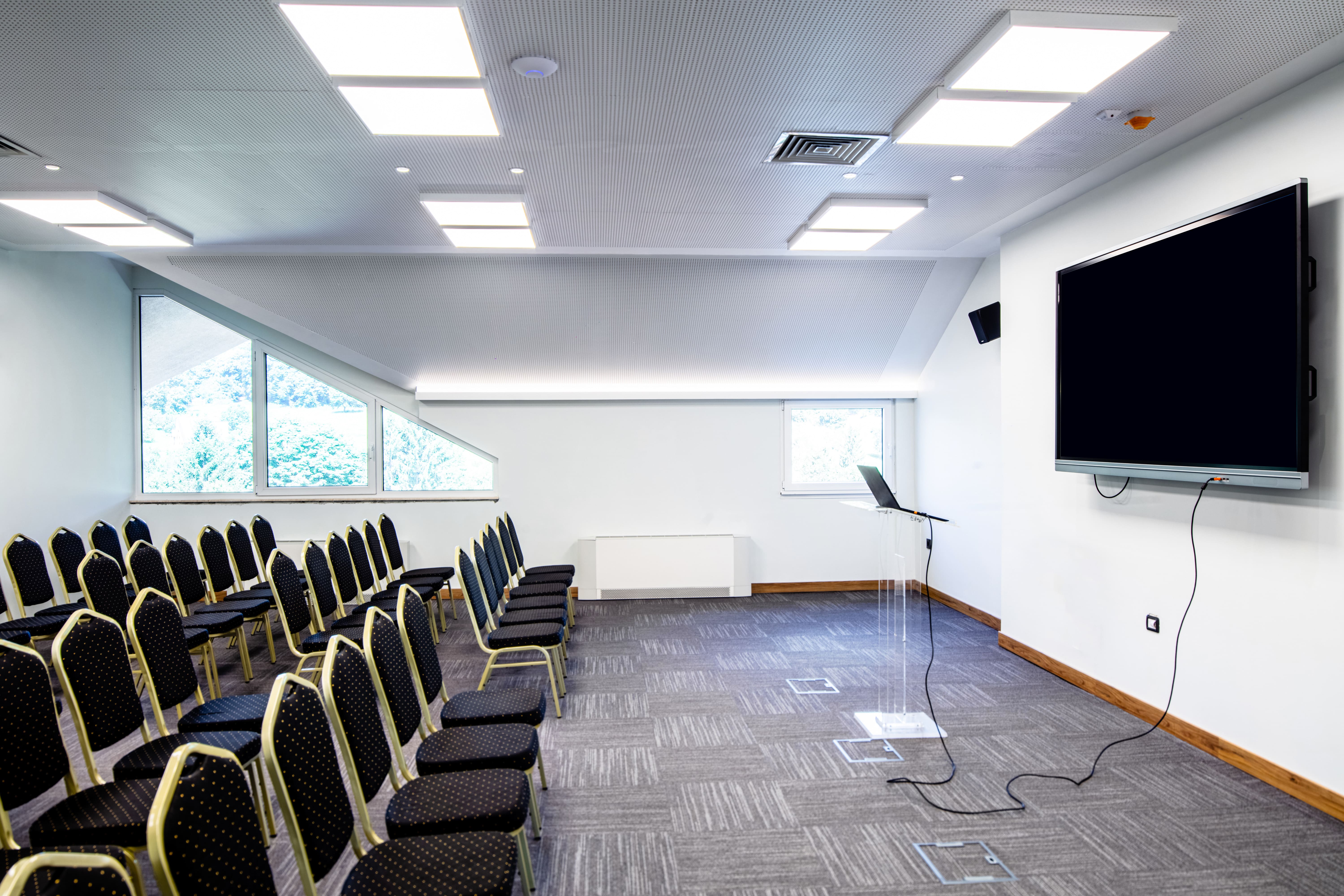 Sala de conferências vazia com cadeiras alinhadas, uma tela de TV na parede e um púlpito com laptop conectado para apresentação.
